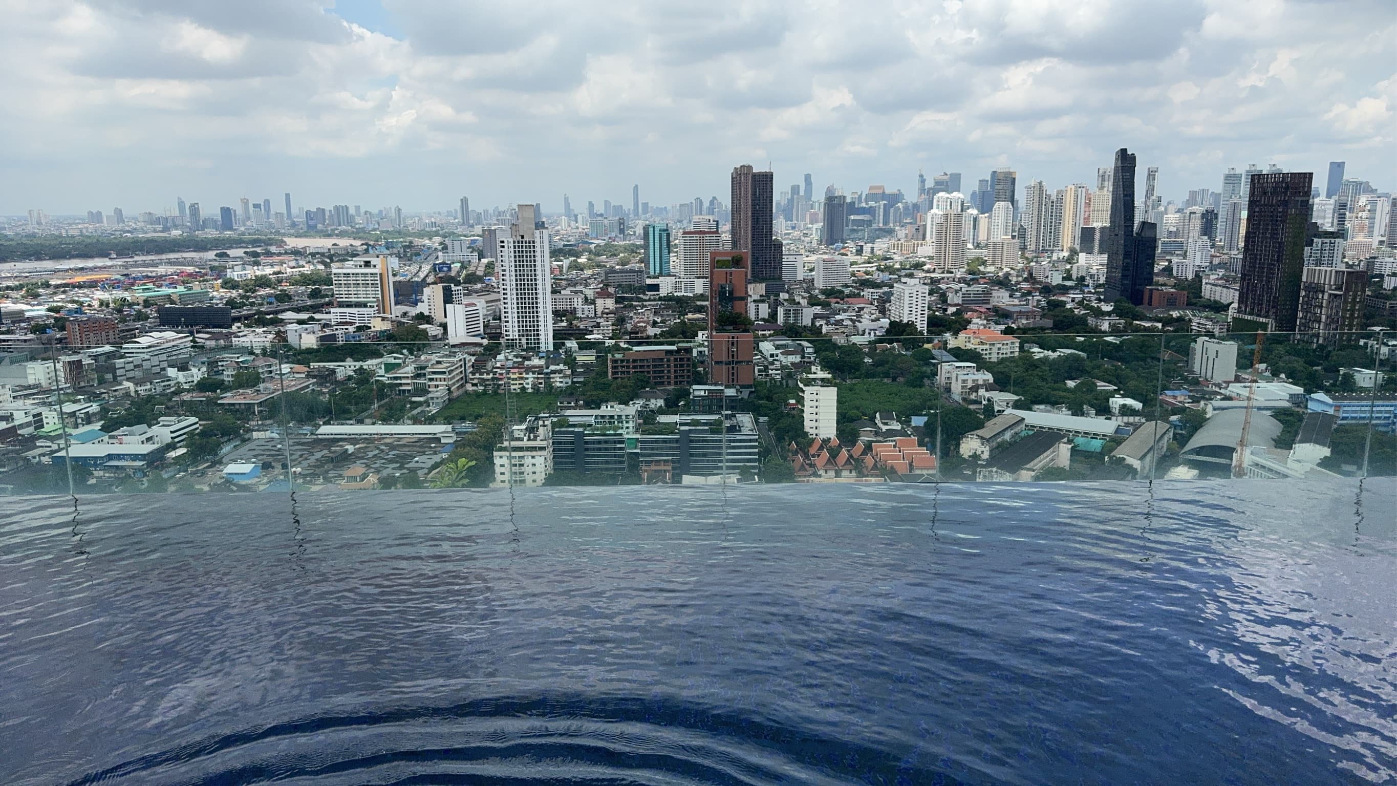 Rooftop infinity pool with water reflecting the Bangkok cityscape, skyscrapers and low-rise buildings stretching to the horizon under scattered clouds