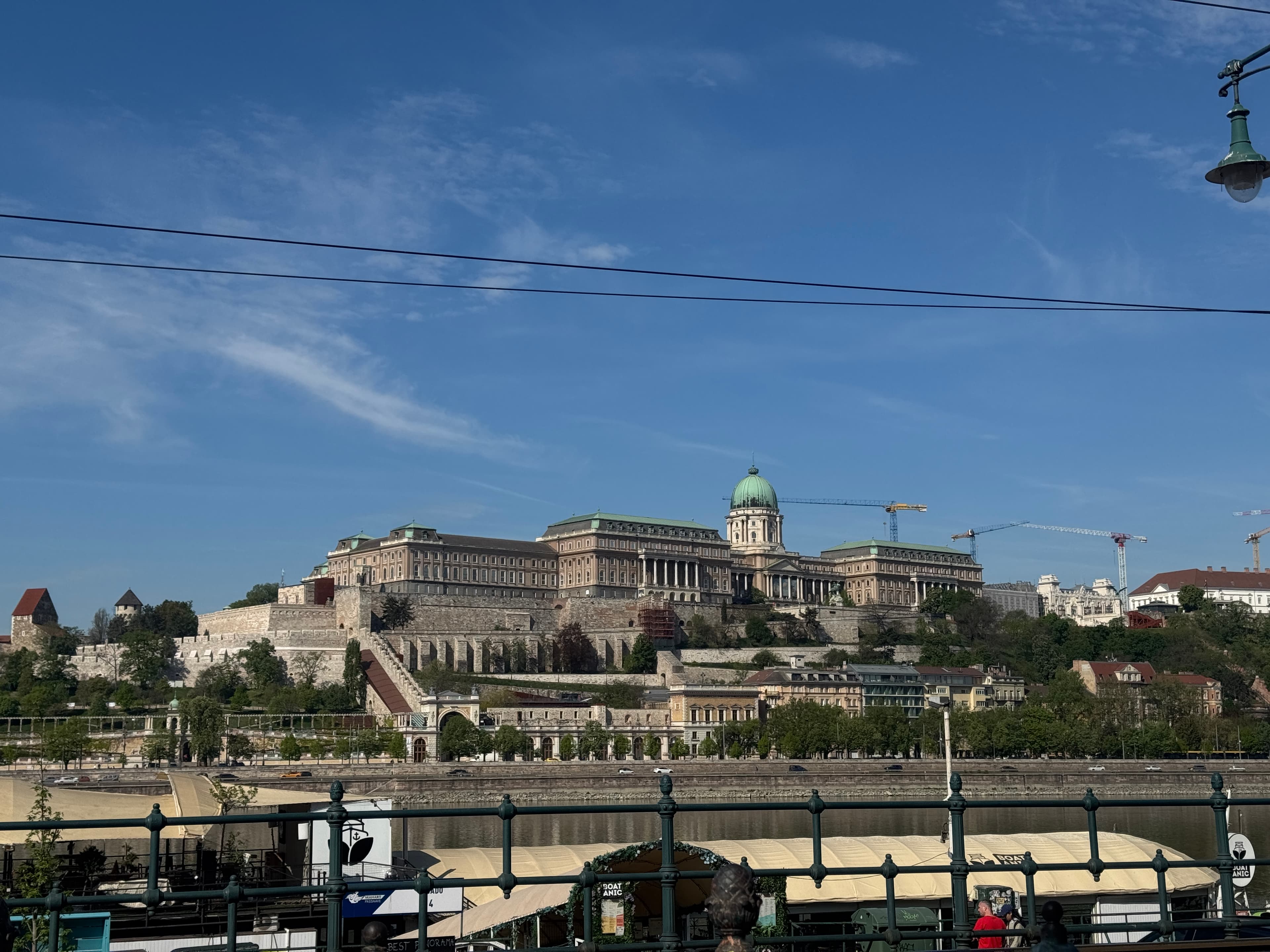 Buda Castle seen across the Danube under a bright blue sky, the green dome of the Royal Palace rising above the fortress walls with construction cranes in the distance