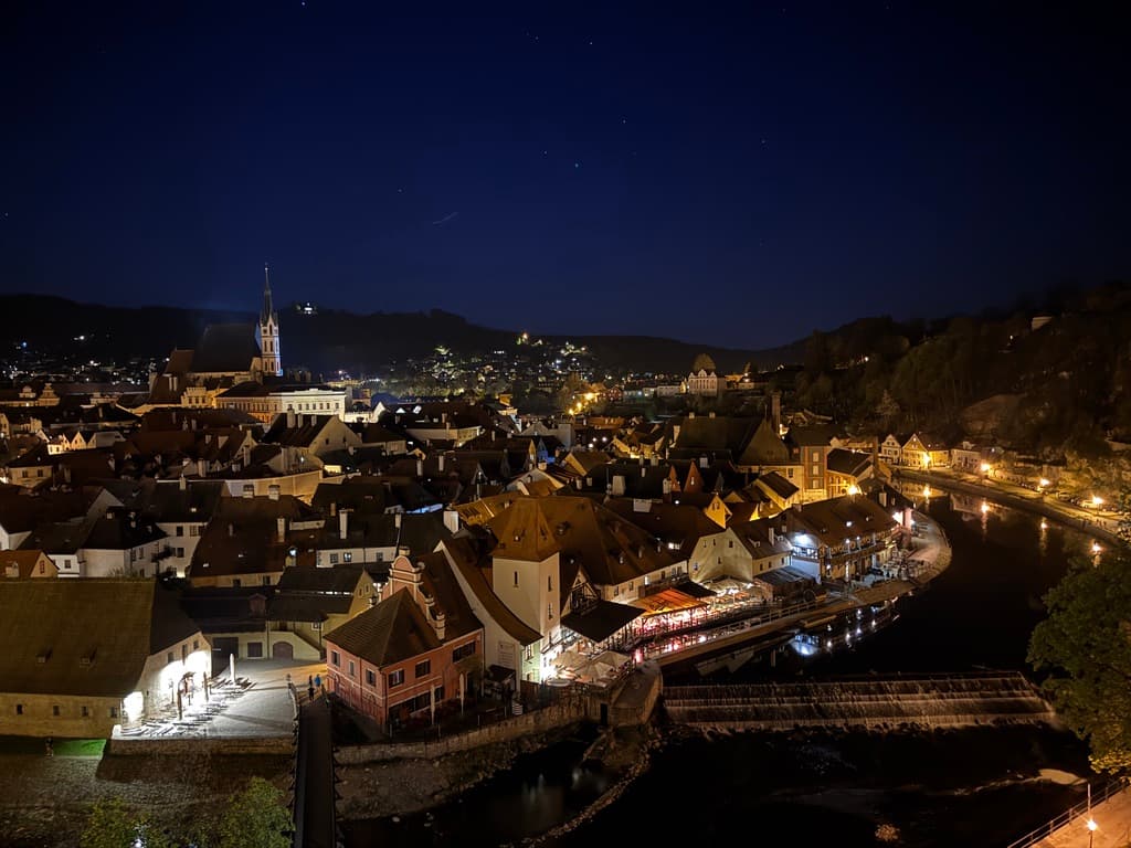 Český Krumlov's old town glowing at night, with St. Vitus Church, clustered red roofs, and the Vltava river beneath a deep blue sky