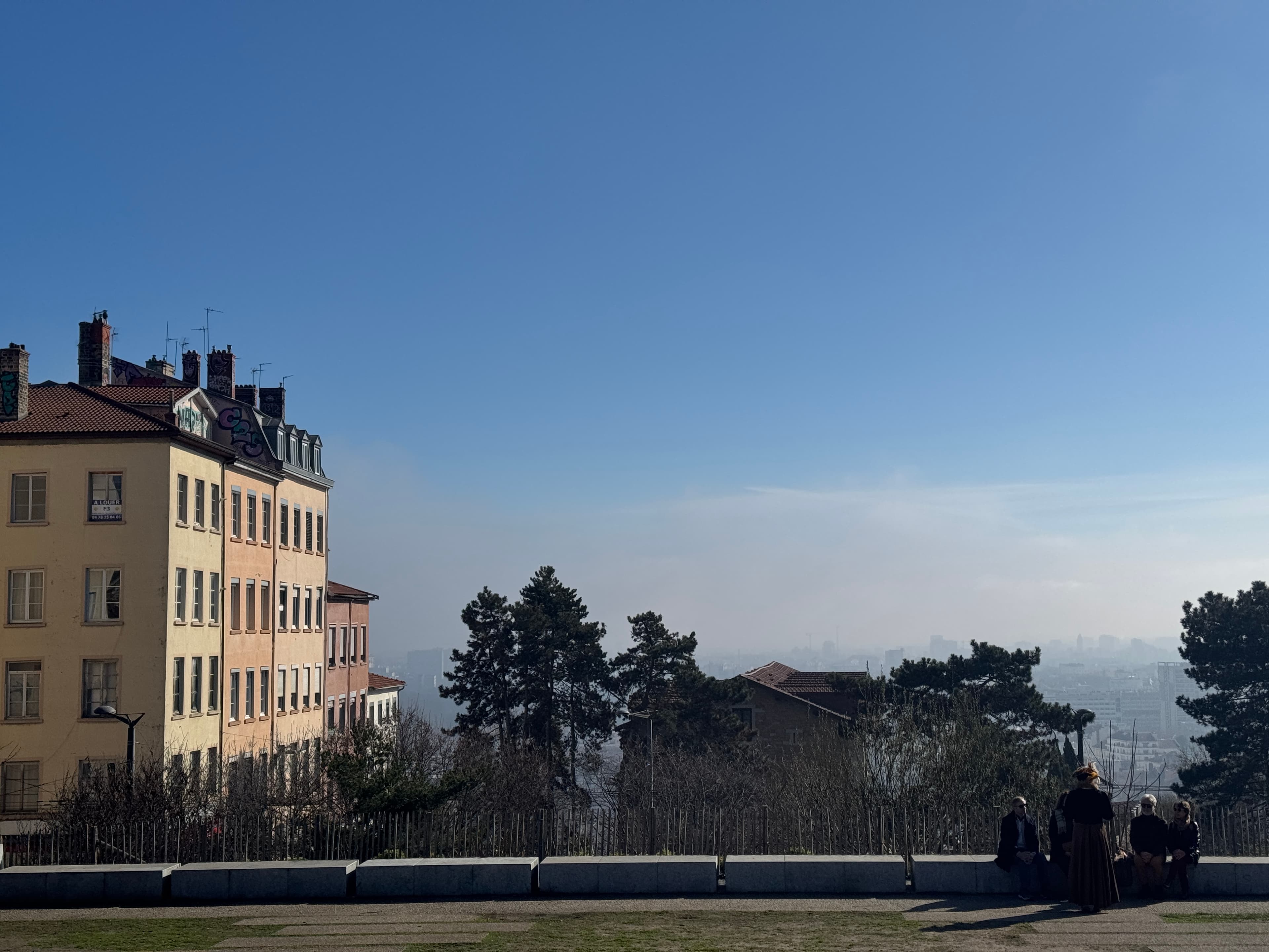 Panoramic view from a Lyon hilltop, city skyline dissolving into winter haze, figures sitting along a low wall