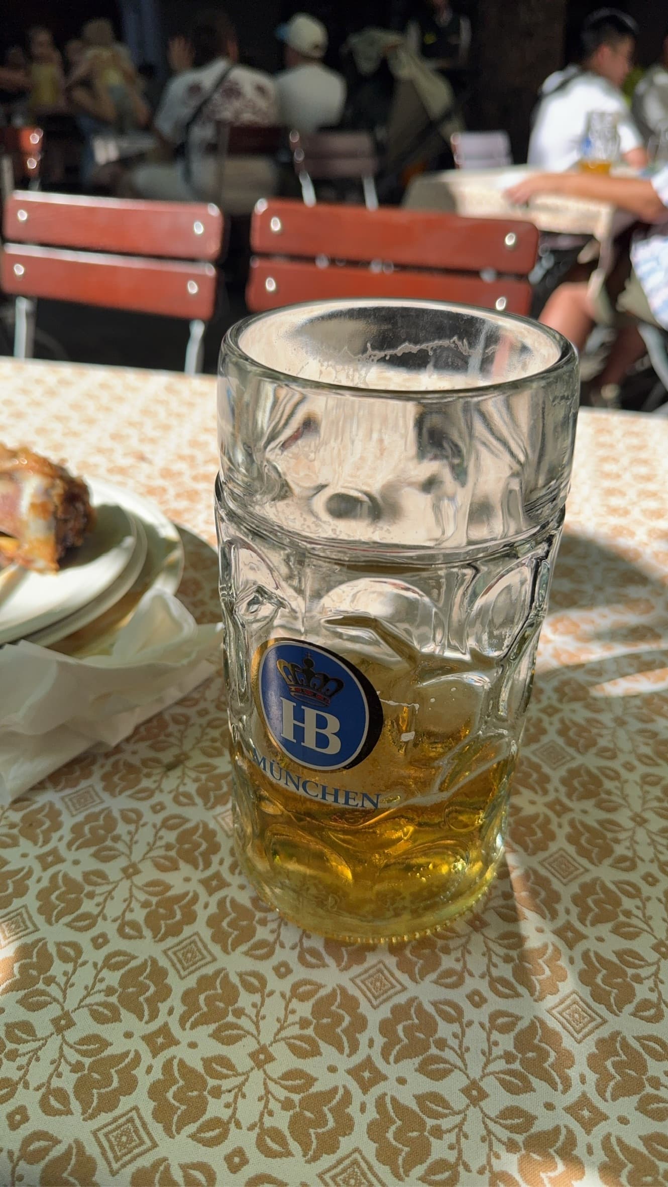 Partly finished Hofbräu beer in a large glass mug on a patterned tablecloth, with a busy Munich beer garden in the background