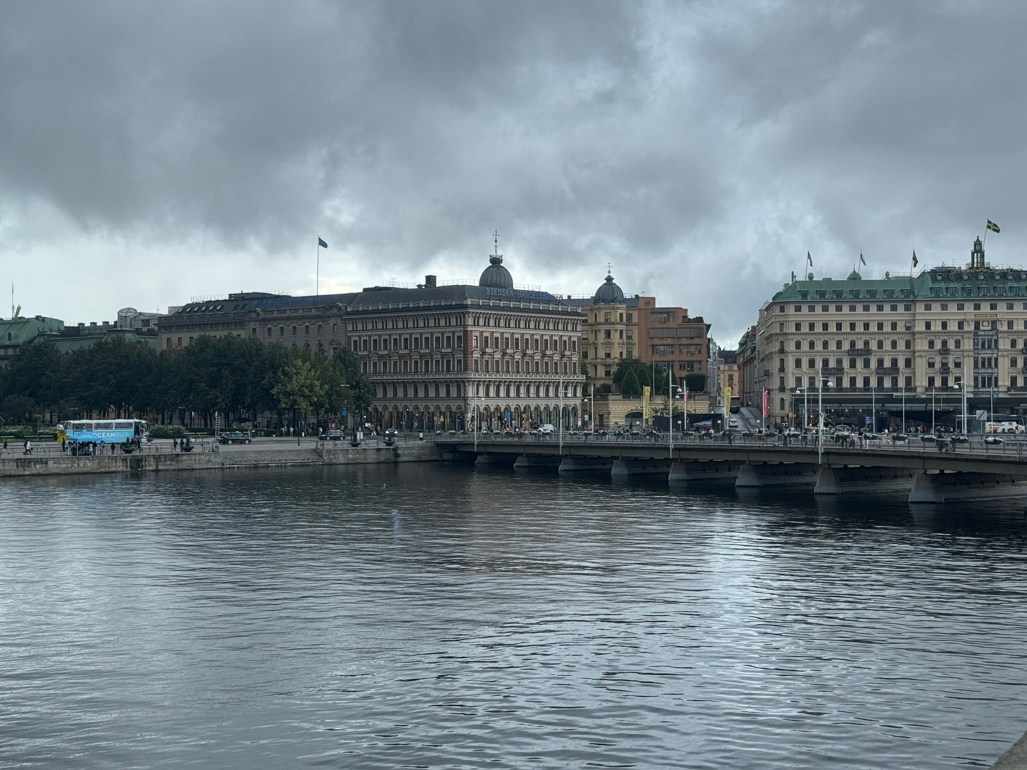 Historic buildings and a busy bridge along Stockholm's waterfront, reflected in calm water beneath heavy grey clouds